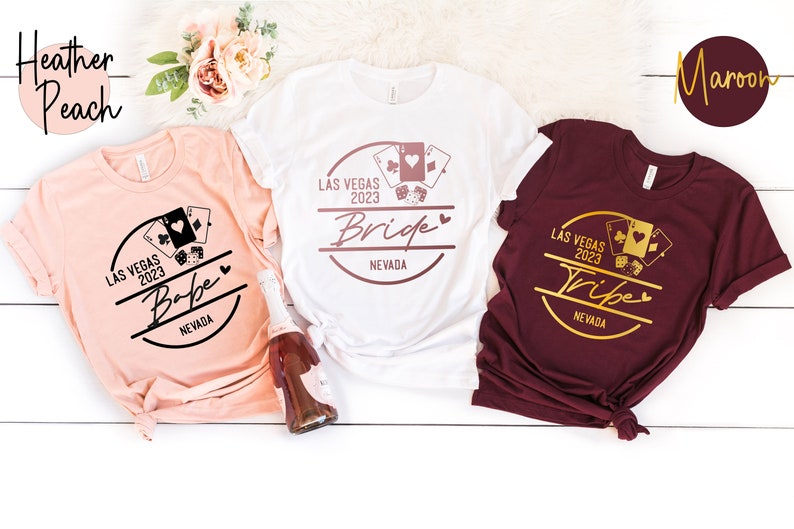 May include: Three t-shirts in heather peach, white, and maroon, each with a Las Vegas 2023 graphic. The heather peach shirt says "Babe", the white shirt says "Bride", and the maroon shirt says "Tribe".