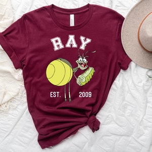 Puede incluir: Camiseta color burdeos con una imagen de dibujos animados de Ray de la pel&iacute;cula de Disney La Princesa y la Rana. La imagen muestra a Ray sosteniendo una luci&eacute;rnaga brillante y el texto "RAY EST. 2009".