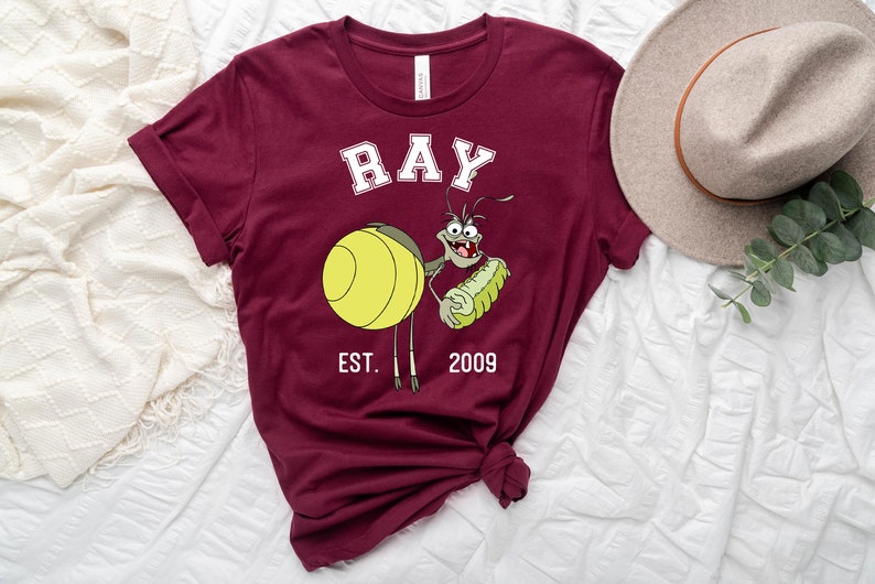 Puede incluir: Camiseta color burdeos con una imagen de dibujos animados de Ray de la pel&iacute;cula de Disney La Princesa y la Rana. La imagen muestra a Ray sosteniendo una luci&eacute;rnaga brillante y el texto "RAY EST. 2009".