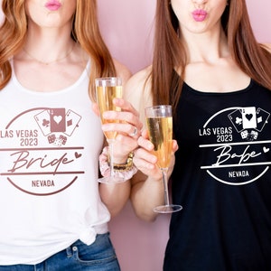 May include: Two women wearing tank tops with a Las Vegas 2023 design. One top is white with "Bride" text, the other is black with "Babe" text. Both tops feature playing card and dice graphics. They are holding champagne flutes.