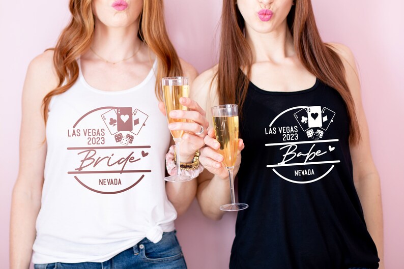 May include: Two women wearing tank tops with a Las Vegas 2023 design. One top is white with "Bride" text, the other is black with "Babe" text. Both tops feature playing card and dice graphics. They are holding champagne flutes.