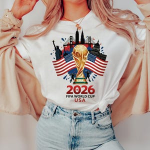 May include: White t-shirt with a gold FIFA World Cup trophy, American flags, and U.S. landmarks. The design includes the text "2026 FIFA WORLD CUP USA" in red and black. A patriotic design.