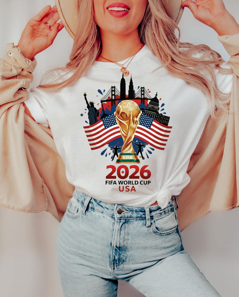 May include: White t-shirt with a gold FIFA World Cup trophy, American flags, and U.S. landmarks. The design includes the text "2026 FIFA WORLD CUP USA" in red and black. A patriotic design.