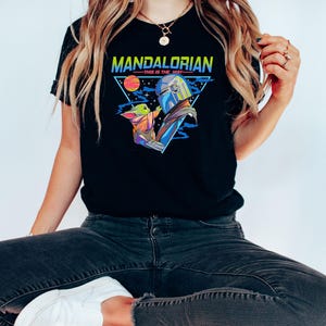 May include: A black t-shirt with a retro-style graphic of a Mandalorian and Grogu. The graphic features a triangle with the words "Mandalorian - This is the Way" in a colorful font.