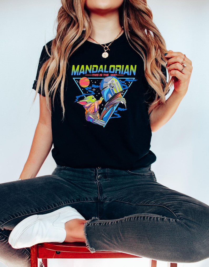 May include: A black t-shirt with a retro-style graphic of a Mandalorian and Grogu. The graphic features a triangle with the words "Mandalorian - This is the Way" in a colorful font.