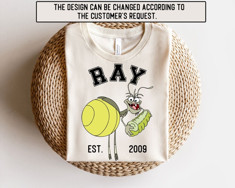 Puede incluir: Una camiseta blanca con una imagen de dibujos animados de Ray de la pel&iacute;cula de Disney, Bichos: Una aventura en miniatura. El texto "RAY" est&aacute; en negro con un contorno negro. El texto "EST. 2009" est&aacute; debajo de la imagen.
