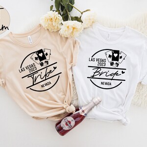 May include: Two t-shirts, one cream and one white, with black graphic designs. The designs include playing cards, dice, and the words "Las Vegas 2023" and "Tribe" or "Bride" with "Nevada" below. A bottle of sparkling wine is in the foreground.