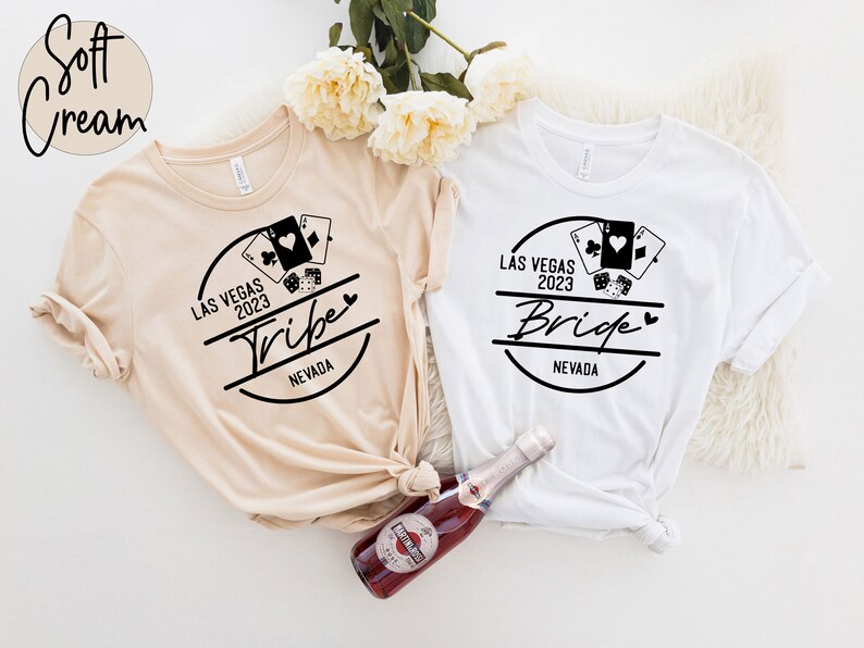 May include: Two t-shirts, one cream and one white, with black graphic designs. The designs include playing cards, dice, and the words "Las Vegas 2023" and "Tribe" or "Bride" with "Nevada" below. A bottle of sparkling wine is in the foreground.
