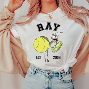 Puede incluir: Camiseta blanca con una imagen de dibujos animados de Ray de La Sirenita de Disney. La imagen muestra a Ray sosteniendo un caballito de mar verde y el texto "RAY EST. 2009".