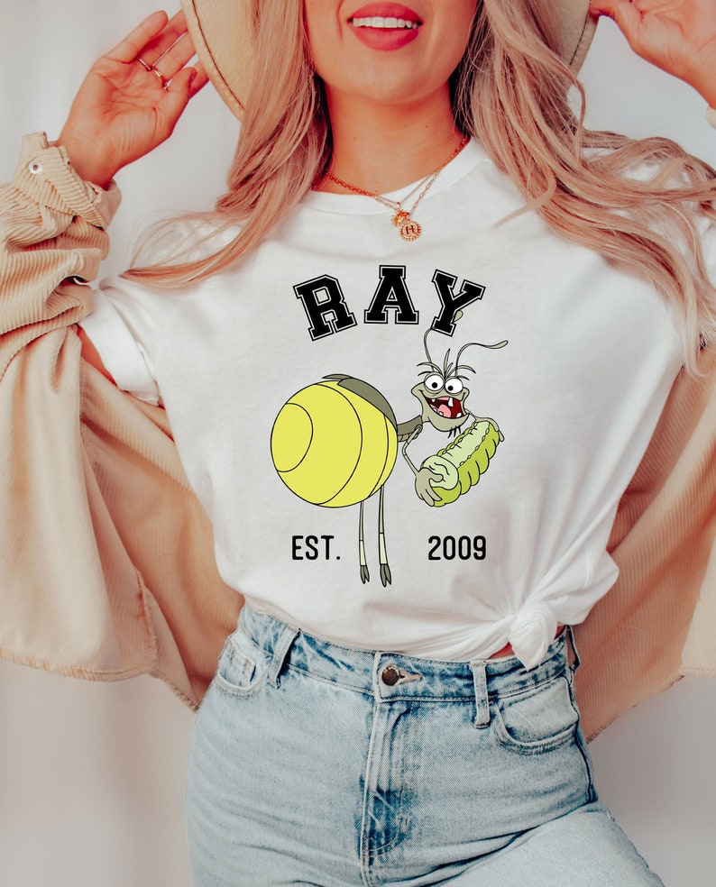 Puede incluir: Camiseta blanca con una imagen de dibujos animados de Ray de La Sirenita de Disney. La imagen muestra a Ray sosteniendo un caballito de mar verde y el texto "RAY EST. 2009".