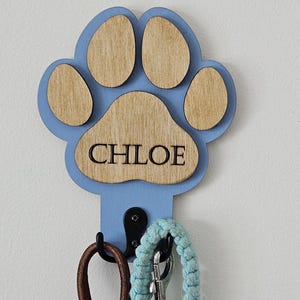 Peut inclure: Un crochet mural en forme d'empreinte de patte bleu clair avec le nom "CHLOE" au centre. Le crochet maintient une laisse en cuir marron et une laisse tressée bleu clair. L'empreinte de patte est en bois et a un bord bleu clair.