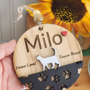 Puede incluir: Un adorno redondo de madera con una silueta de gato y un dise&ntilde;o de huellas de patas. El adorno est&aacute; personalizado con el nombre "Milo" y el texto "Forever Loved" y "Forever Missed".