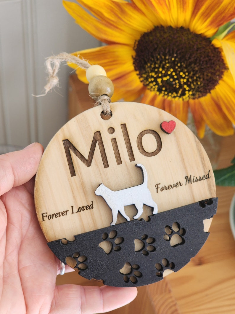 Puede incluir: Un adorno redondo de madera con una silueta de gato y un dise&ntilde;o de huellas de patas. El adorno est&aacute; personalizado con el nombre "Milo" y el texto "Forever Loved" y "Forever Missed".