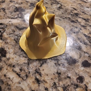 Puede incluir: Un modelo de sombrero de mago impreso en 3D de color dorado, con una punta puntiaguda y un ala ancha.