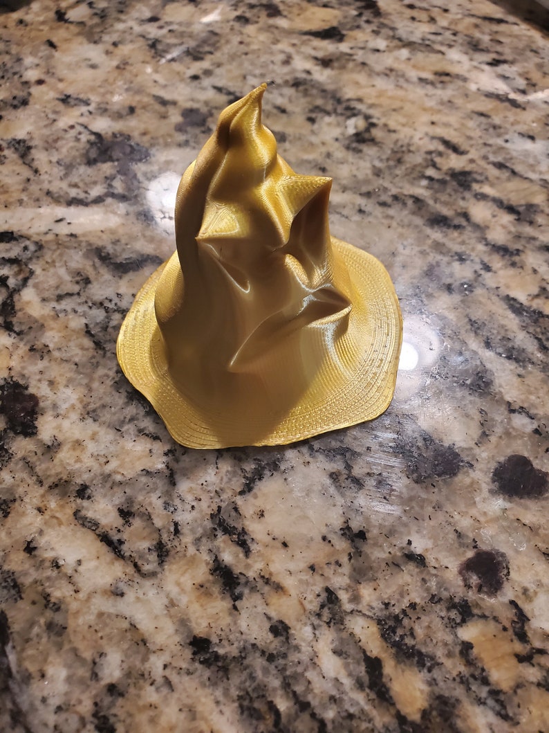 Puede incluir: Un modelo de sombrero de mago impreso en 3D de color dorado, con una punta puntiaguda y un ala ancha.