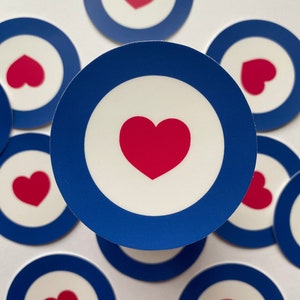May include: A set of eight blue and white target-style stickers with a red heart in the center of each. The stickers are arranged in a circle on a white background.