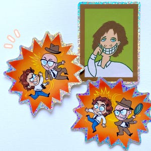 Peut inclure: Trois autocollants colorés représentant des personnages de dessins animés. Le premier autocollant montre deux hommes en train de se battre, le deuxième autocollant montre un homme avec un grand sourire et le troisième autocollant montre deux hommes avec une loupe.