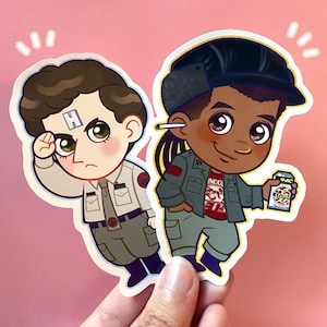 May include: Two cartoon character stickers, one wearing a green and brown uniform and the other wearing a green and red uniform. The character in the green and brown uniform is saluting and has a white letter H on their forehead. The character in the green and red uniform is holding a small box with the text "IMC LEOPARD" on it.