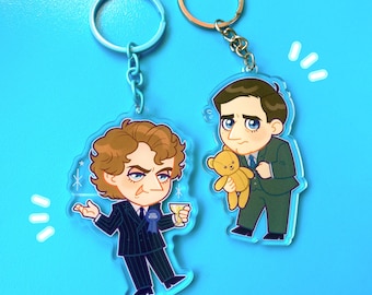May include: Two acrylic keychains featuring cartoon characters. One keychain depicts a character in a pinstripe suit holding a drink, the other shows a character in a green suit holding a teddy bear. Both keychains have metal rings.