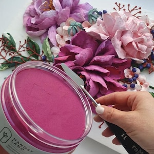 Peut inclure: Gros plan sur une peinture acrylique rose dans un pot transparent avec une étiquette blanche qui dit "HANDSY LANDS". Une personne utilise une petite spatule en métal pour prélever de la peinture.