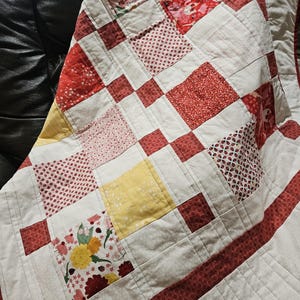May include: A patchwork quilt featuring a grid of square fabric blocks in shades of red, white, and yellow. The design includes various patterns such as floral prints, small dots, and stripes, all stitched together with visible quilting lines.