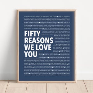 May include: A navy blue poster with white text that reads "Fifty Reasons We Love You". The poster is framed in a light wood frame and is hanging on a white wall.