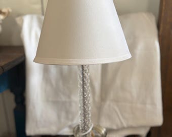 May include: A clear glass table lamp with a white lampshade. The lamp has a textured base and a beaded stem. The lampshade is a light cream color and is cone-shaped.