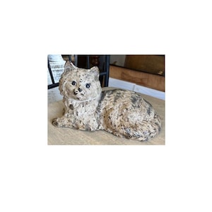 May include: A detailed, textured cat figurine in a resting pose. The cat is primarily beige with darker spots and a small collar. The sculpture has a vintage or antique appearance.