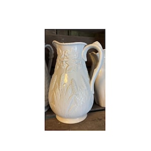 May include: White ceramic jug with an embossed design of wheat and flowers. The jug has a wide mouth and a handle.