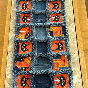 Puede incluir: Un camino de mesa de mezclilla con un diseño de patchwork. El camino de mesa presenta cuadrados de mezclilla azul oscuro y tela naranja con caras de gatos negros. Los bordes de los cuadrados de mezclilla están deshilachados.