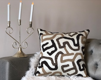 May include: A white throw pillow with a brown and black geometric pattern. The pillow is sitting on a white faux fur throw and a gray couch.