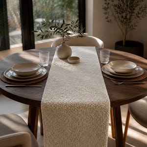 May include: A wooden dining table set with a cream-coloured, textured table runner. Two place settings with plates, bowls, and cutlery are arranged on woven placemats. A small vase with greenery and glasses complete the setting. The table is round.