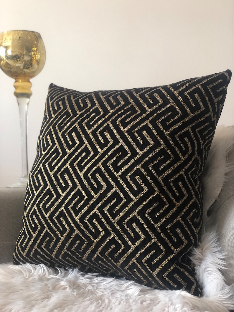 May include: Black and gold decorative throw pillow with a geometric pattern. The pillow is made of a soft, plush fabric and features a repeating pattern of interlocking squares and rectangles.