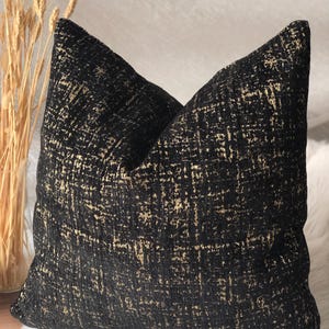 Peut inclure: Un coussin carré décoratif avec un motif texturé noir et or. Le coussin a un aspect tissé avec un motif quadrillé. Le coussin est posé sur une surface blanche et moelleuse, avec un vase de tiges de blé séchées en arrière-plan.