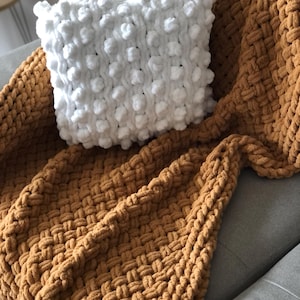 May include: A white knitted throw pillow with a textured bobble design sits on top of a brown knitted throw blanket.