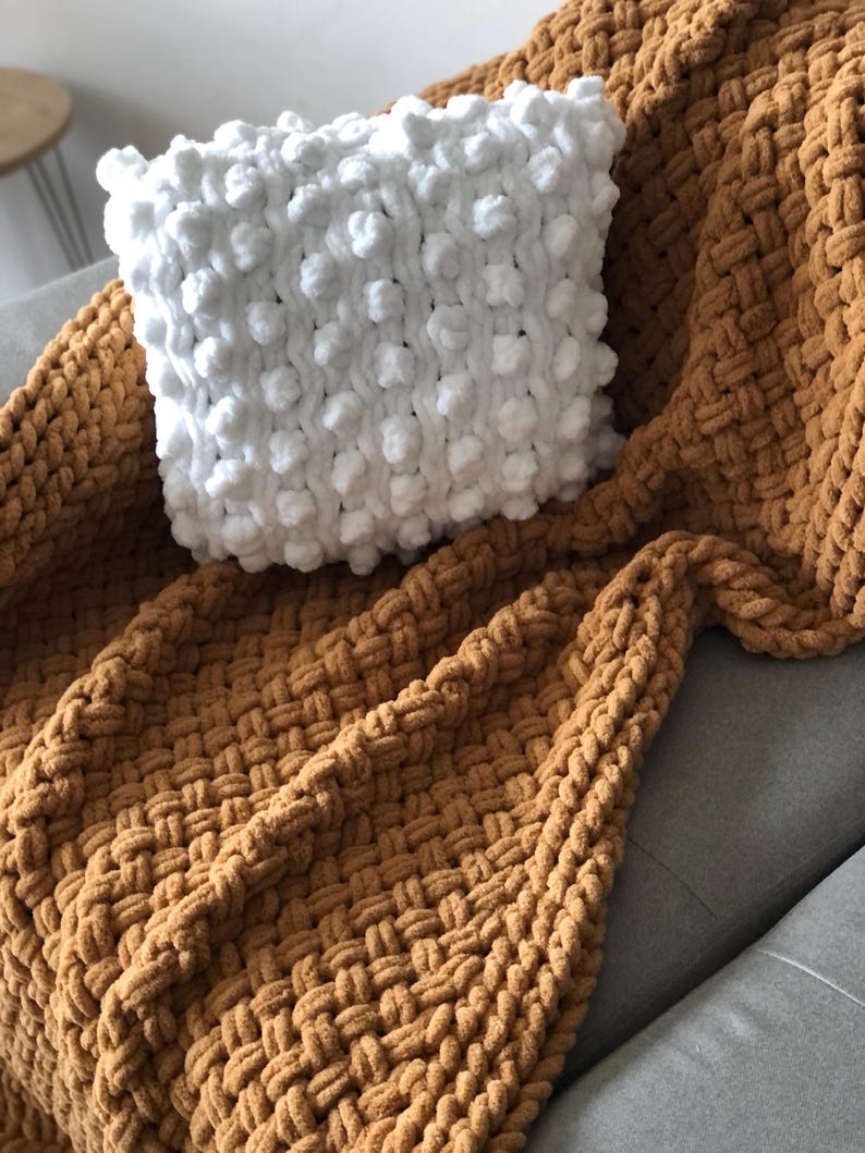 May include: A white knitted throw pillow with a textured bobble design sits on top of a brown knitted throw blanket.
