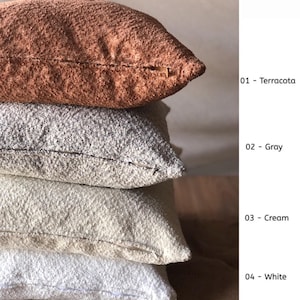 May include: A stack of four square throw pillows with a textured, nubby fabric surface. The colors from top to bottom are labeled as 01 - Terracota, 02 - Gray, 03 - Cream, and 04 - White. Each pillow features a visible side seam and zipper closure.