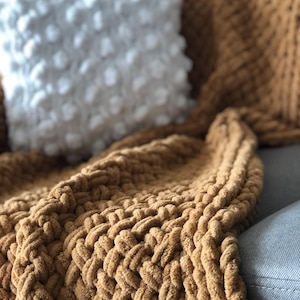 May include: A brown knitted throw blanket with a chunky cable knit pattern. The blanket is draped over a couch with a white pillow.