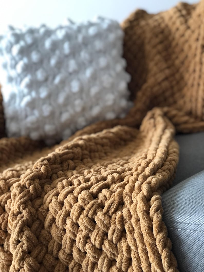 May include: A brown knitted throw blanket with a chunky cable knit pattern. The blanket is draped over a couch with a white pillow.
