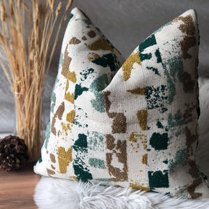 May include: A white throw pillow with a geometric pattern in shades of green, brown, and yellow. The pillow is made of a textured fabric and has a square shape.