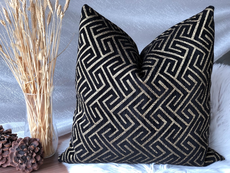 May include: Black decorative pillow with a gold geometric pattern. The pillow is a square shape and has a soft, textured appearance. The pillow is placed on a white surface. Decorative home decor.