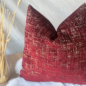 May include: A decorative throw pillow with a textured, red fabric and gold accents. The pillow has a square shape and is placed on a white, fluffy surface. A vase of dried wheat stalks is visible in the background.