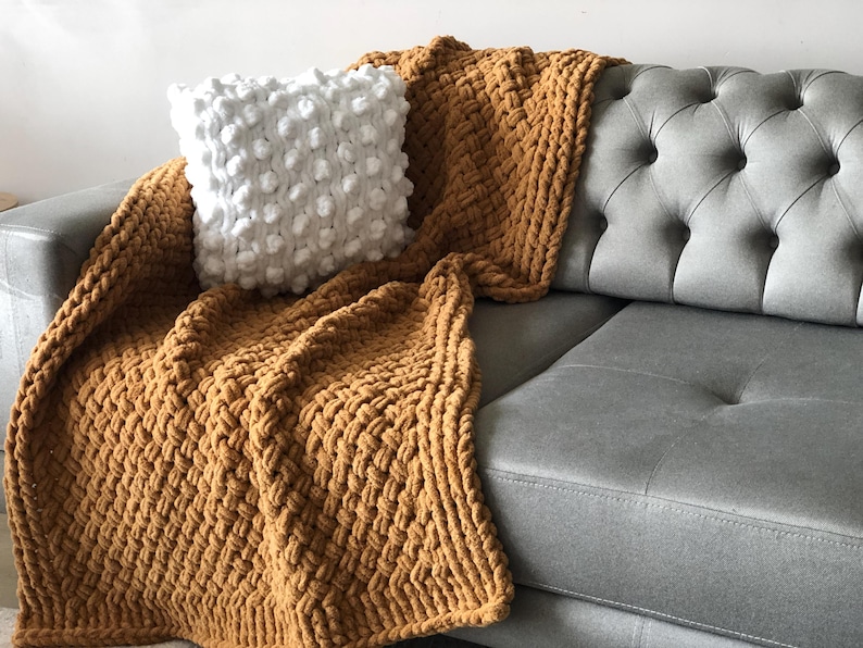 May include: A mustard yellow chunky knit throw blanket draped over a gray tufted sofa with a white textured throw pillow.