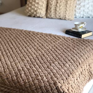 May include: A brown knitted throw blanket with a textured waffle weave pattern. The blanket is draped over a white bed with decorative cushions.