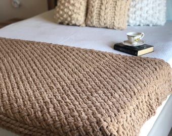 May include: A brown knitted throw blanket with a textured waffle weave pattern. The blanket is draped over a white bed with decorative pillows.