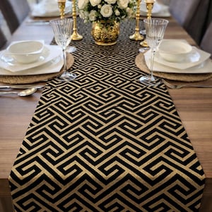 Könnte beinhalten: Ein Esstisch für einen formellen Anlass. Der Tisch ist mit einem schwarz-goldenen Tischläufer mit geometrischem Muster, weißen Tellern und Kristallgläsern geschmückt. Goldene Kerzenhalter und ein Blumengesteck vervollständigen die elegante Einrichtung.