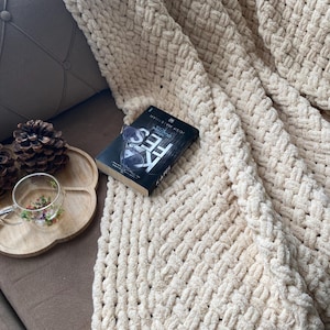 May include: A cream-colored, chunky knit blanket draped over a brown surface. A book with the title "FES" is on the blanket. A wooden tray with a glass teacup and pinecones is visible.