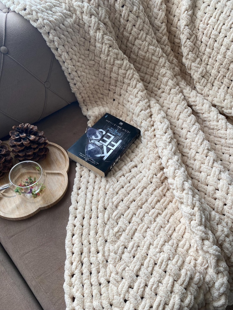 May include: A cream-colored, chunky knit blanket draped over a brown surface. A book with the title "FES" is on the blanket. A wooden tray with a glass teacup and pinecones is visible.