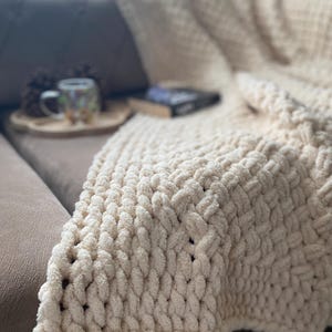 May include: A cream-colored, chunky knit blanket draped over a brown sofa. The blanket has a thick, textured weave. A wooden tray with a mug and a book are visible on the sofa. The blanket appears soft and cozy.