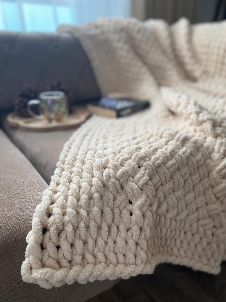 May include: A cream-colored, chunky knit blanket draped over a brown sofa. The blanket has a thick, textured weave. A wooden tray with a mug and a book are visible on the sofa. The blanket appears soft and cozy.